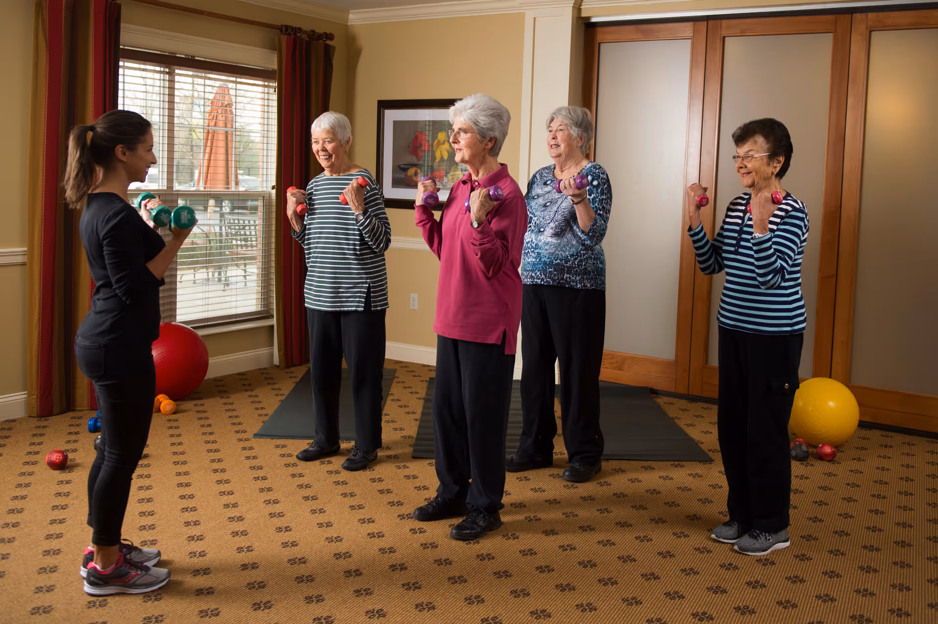 A fitness instructor leads a group exercise session with four elderly women in a carpeted room. Each participant is holding small dumbbells and following the instructor's movements. The room has large windows with curtains, framed artwork on the wall, and exercise mats and balls in the background.