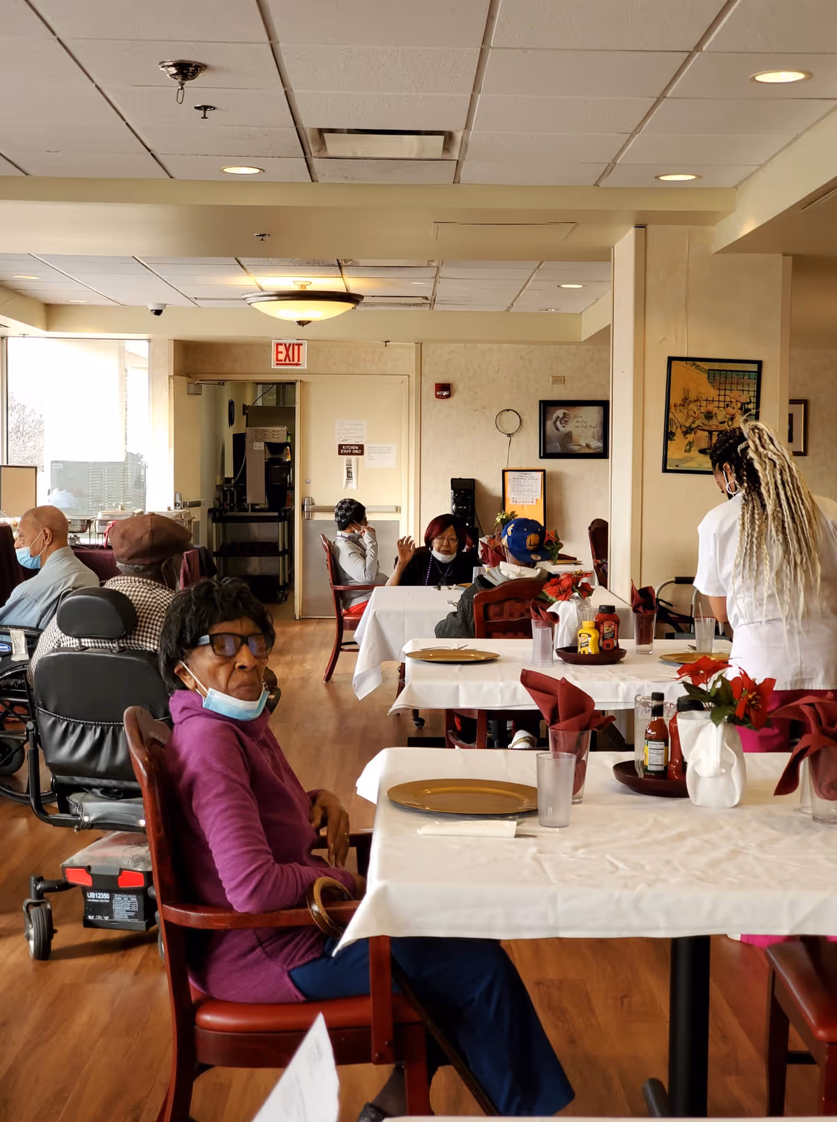 A dining room in a senior living facility with several elderly residents seated at tables covered with white tablecloths and red napkins. A staff member with long braided hair is attending to one of the tables. The room has wooden floors, framed artwork on the walls, and an exit door in the background.