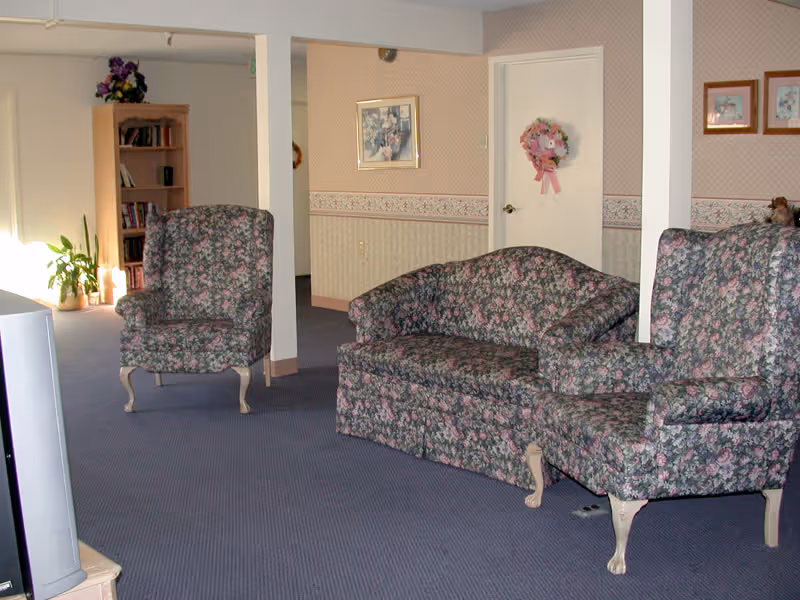 A living room area with floral patterned furniture including a loveseat and two armchairs. The room has blue carpet, wallpaper with a floral border, framed pictures on the walls, and a door decorated with a pink floral wreath. A bookshelf and a potted plant are visible in the background.