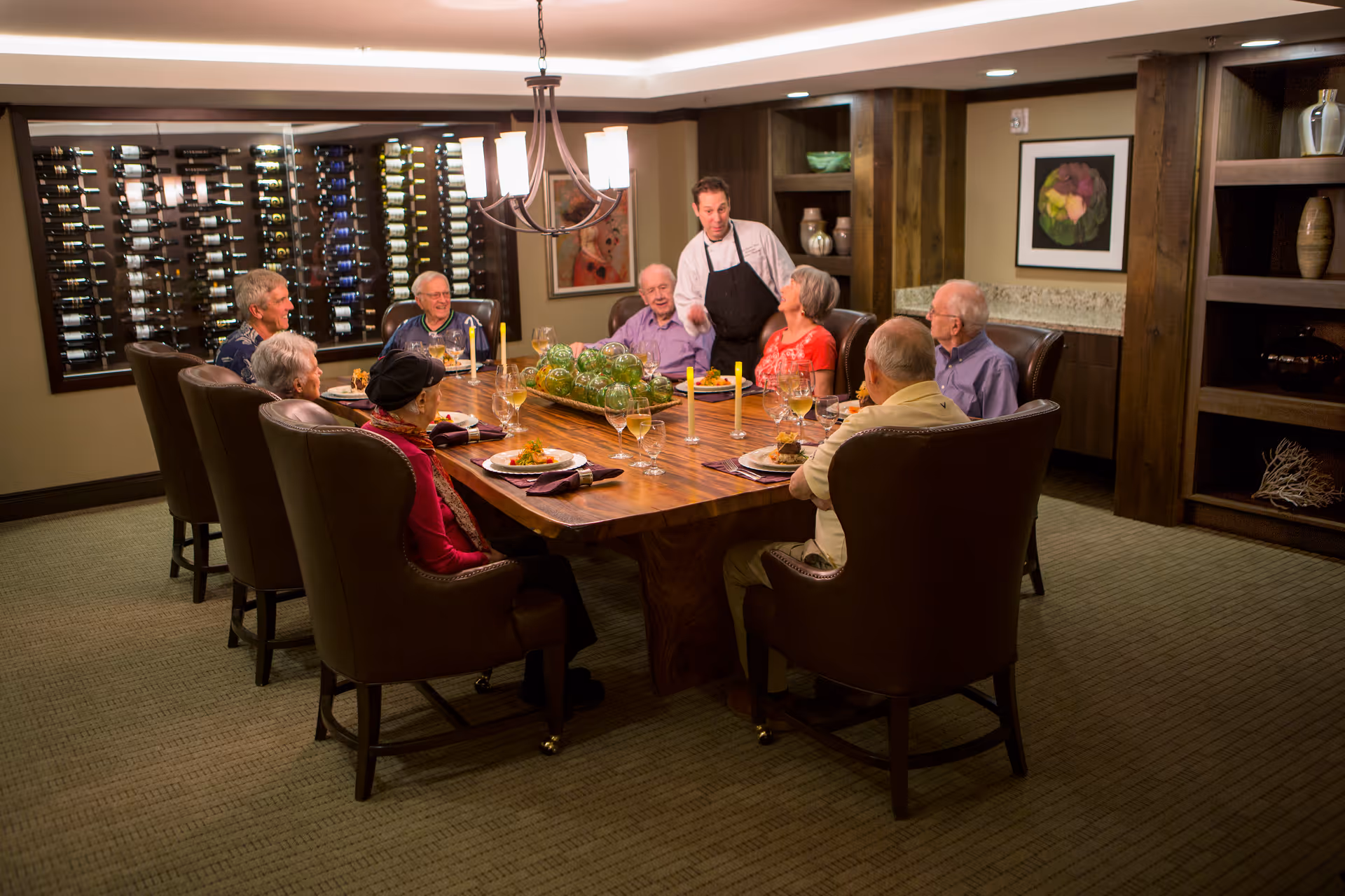 A group of elderly people seated around a large wooden dining table in a warmly lit room, with a waiter standing and engaging with them. The table is set with plates of food, wine glasses, and candles. The background features a wall-mounted wine rack and wooden shelves with decorative items.