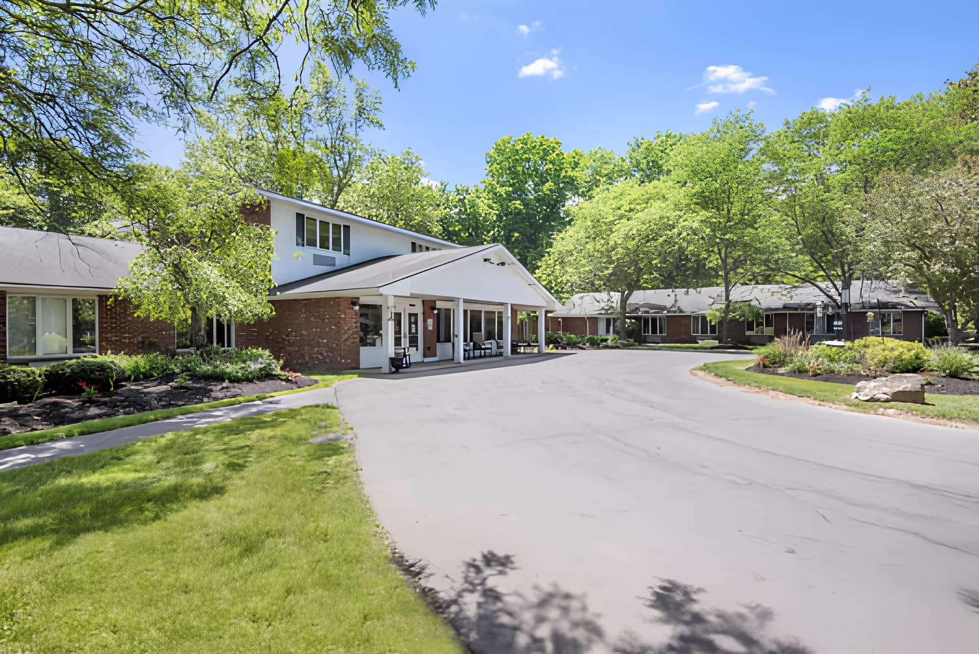 Exterior view of Brookdale East Side facility showing a driveway leading to the entrance with a covered porch. The building is surrounded by green trees and well-maintained landscaping under a clear blue sky.