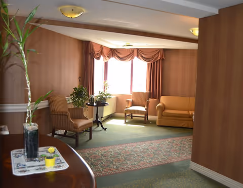A cozy sitting area in an assisted living facility with two upholstered armchairs, a small round table with plants, a yellow sofa, and large windows with draped curtains letting in natural light. The walls are covered with brown striped wallpaper, and there is a patterned carpet on the floor.