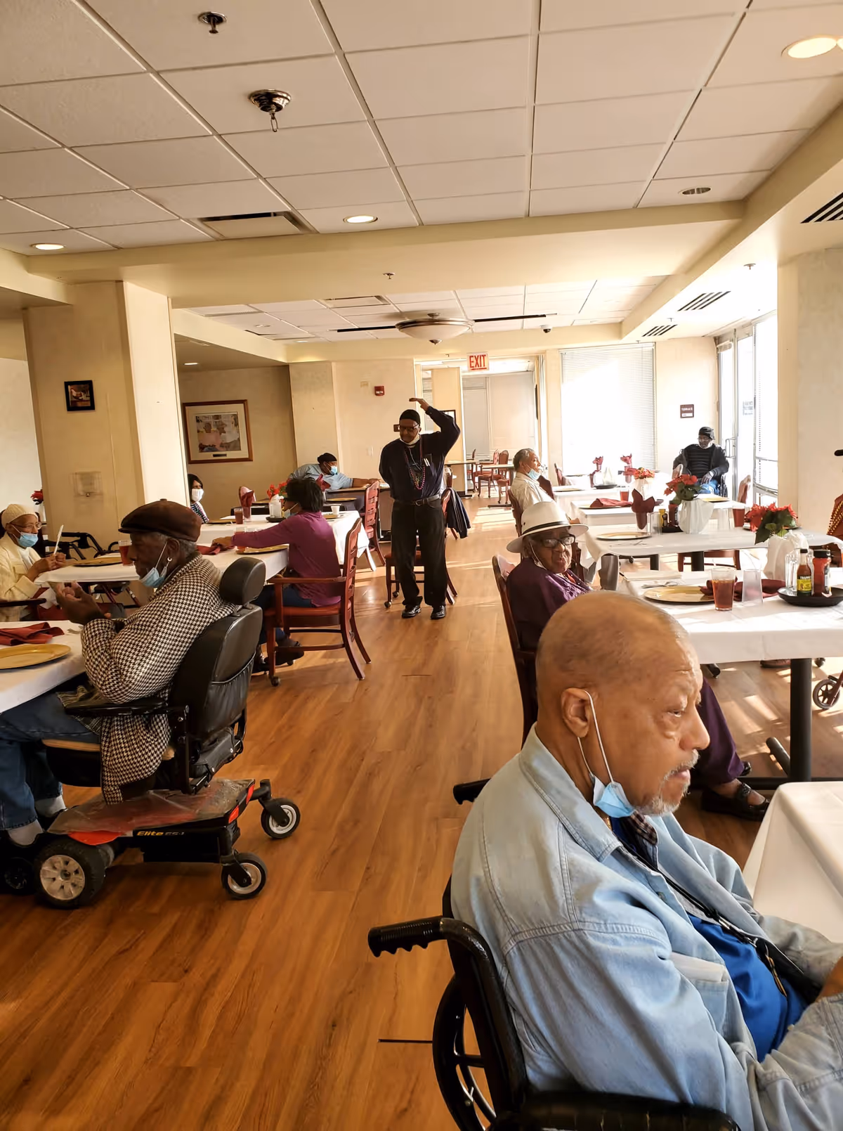 A dining room in a senior living facility with several elderly residents seated at tables. Some are in wheelchairs or mobility scooters, and a few are wearing face masks. The room has wooden flooring, white walls, and large windows letting in natural light. Tables are set with white tablecloths, red napkins, and some have flower centerpieces.