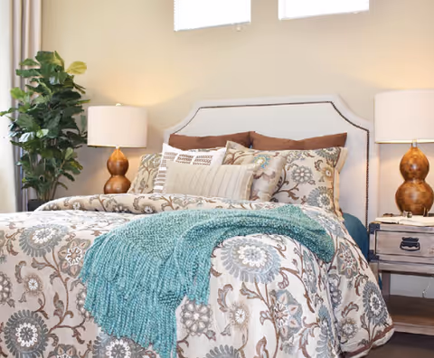 A cozy bedroom featuring a bed with a patterned floral comforter, multiple decorative pillows, and a teal throw blanket. There are two matching bedside tables with brown lamps and a large green plant in the corner near a window with blinds.