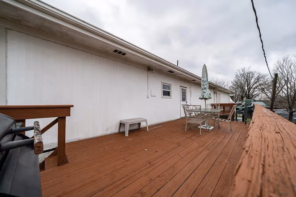 Outdoor wooden deck attached to a building with a white exterior wall. The deck has a glass table with a closed umbrella and metal chairs, a small plastic bench, and some stacked plastic chairs. The sky is cloudy and there are leafless trees in the background.