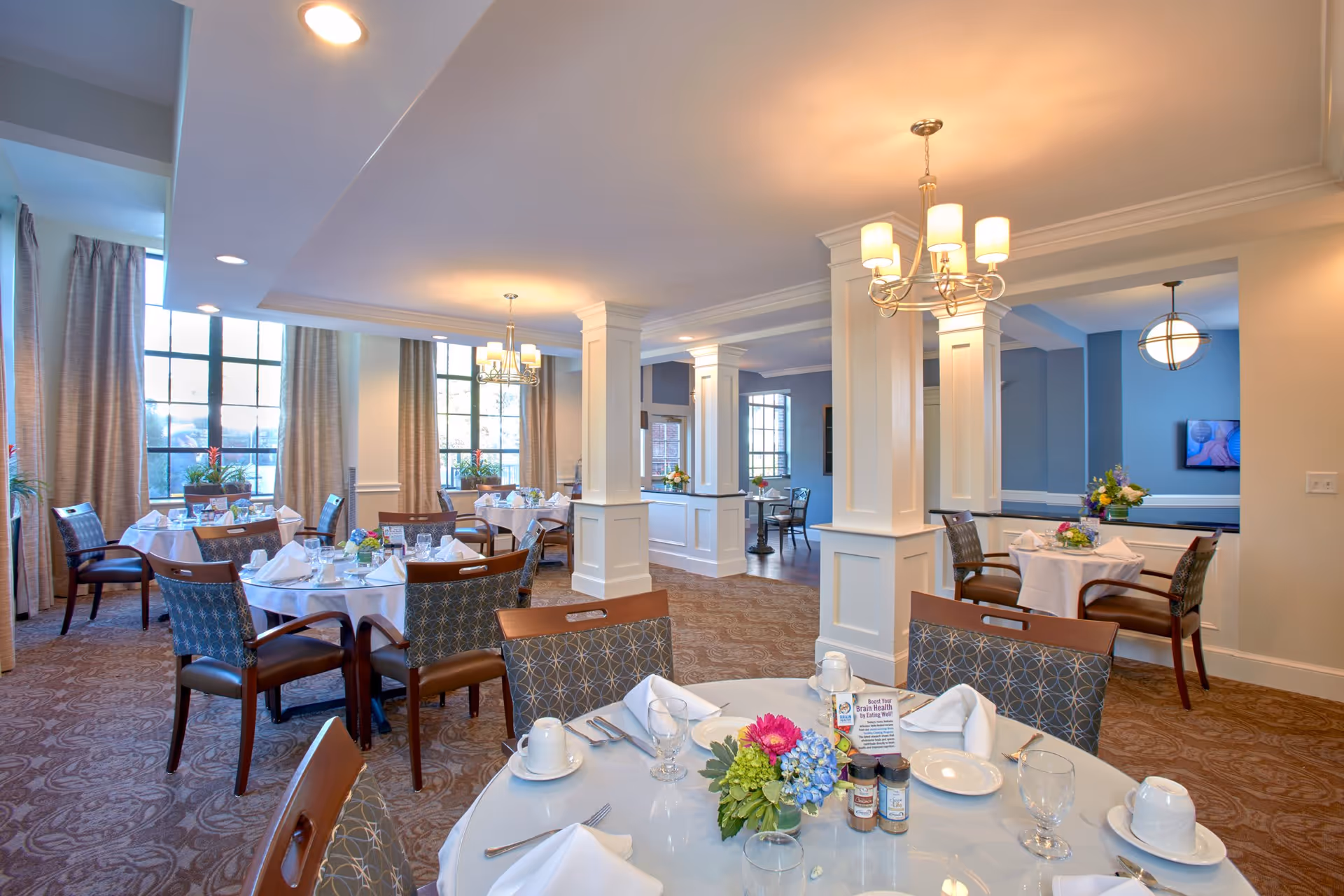 A bright and elegant dining room with round tables covered in white tablecloths, set with plates, glasses, napkins, and silverware. Each table has a floral centerpiece and condiments. The room features large windows with beige curtains, carpeted floors, white columns, and warm overhead lighting fixtures.