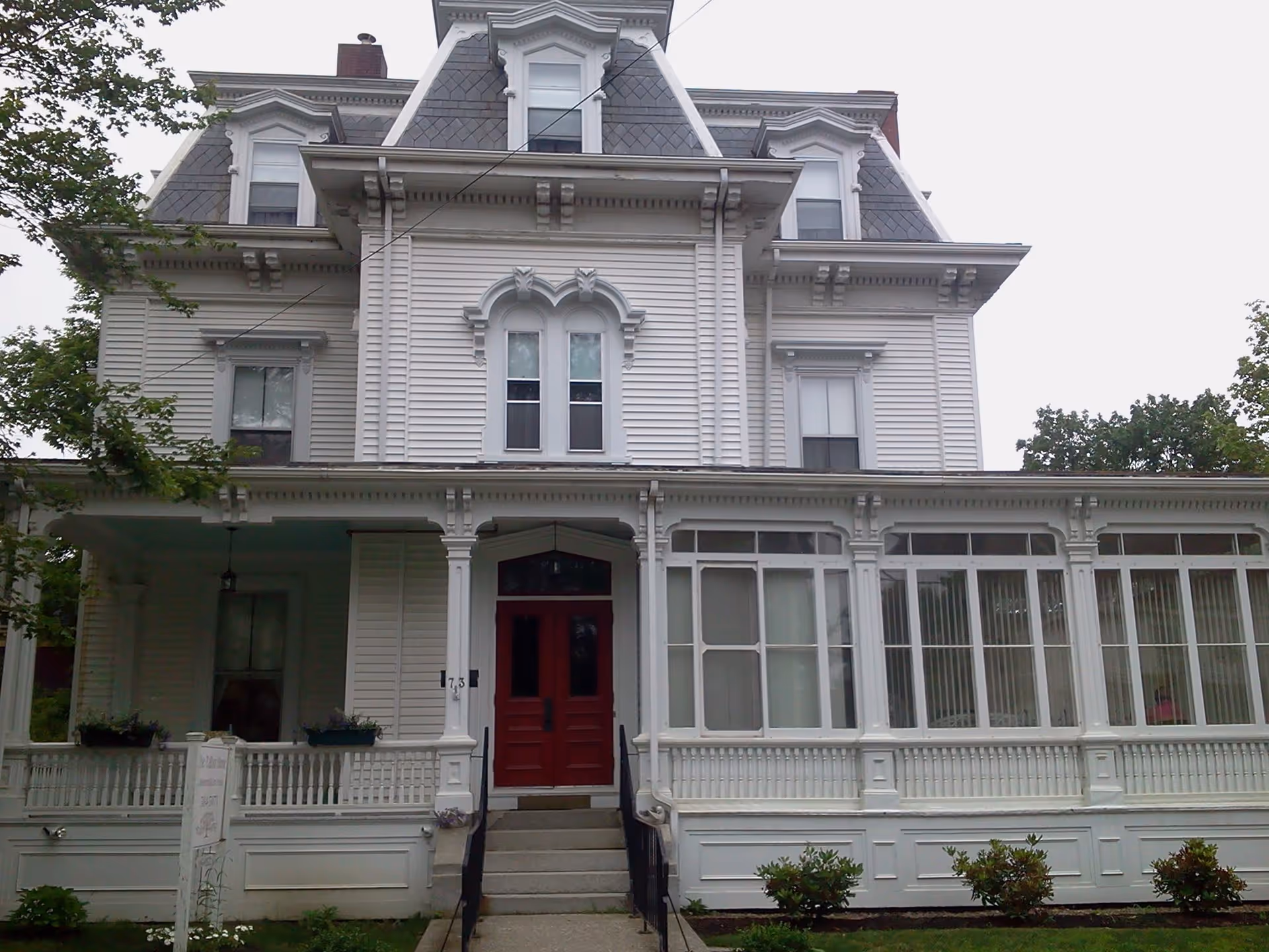Front exterior view of a large, white, Victorian-style house with a red double door entrance, multiple windows, and a covered porch with decorative trim. There are small bushes and a lawn in front of the building.