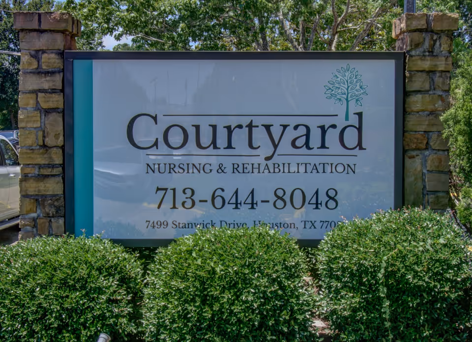 Outdoor sign for Courtyard Nursing & Rehabilitation with phone number 713-644-8048 and address 7499 Stanwick Drive, Houston, TX 77099, surrounded by green bushes and stone pillars.