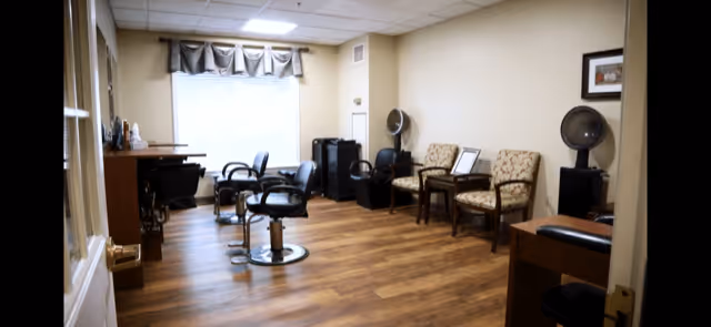 Interior view of a salon area in a senior living facility with wooden flooring, salon chairs, hair dryers, and a window with a valance. There are also a couple of upholstered chairs along the wall and a small table.