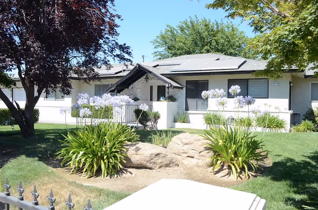 Single-story building with white exterior walls and a gray roof, surrounded by green grass, flowering plants, and trees under a clear blue sky.