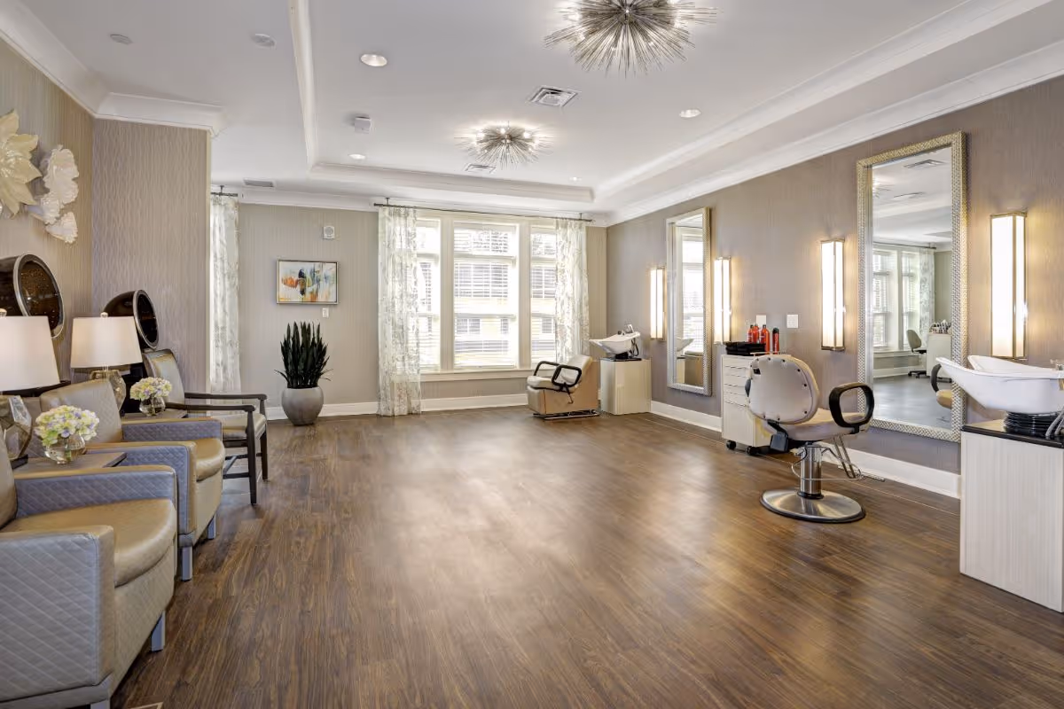 A spacious and well-lit hair salon area with wooden flooring, two styling chairs in front of large mirrors with wall-mounted lights, a hair washing station, and comfortable seating with armchairs and side tables on the left side. The room has large windows with sheer curtains, decorative wall art, and modern ceiling light fixtures.