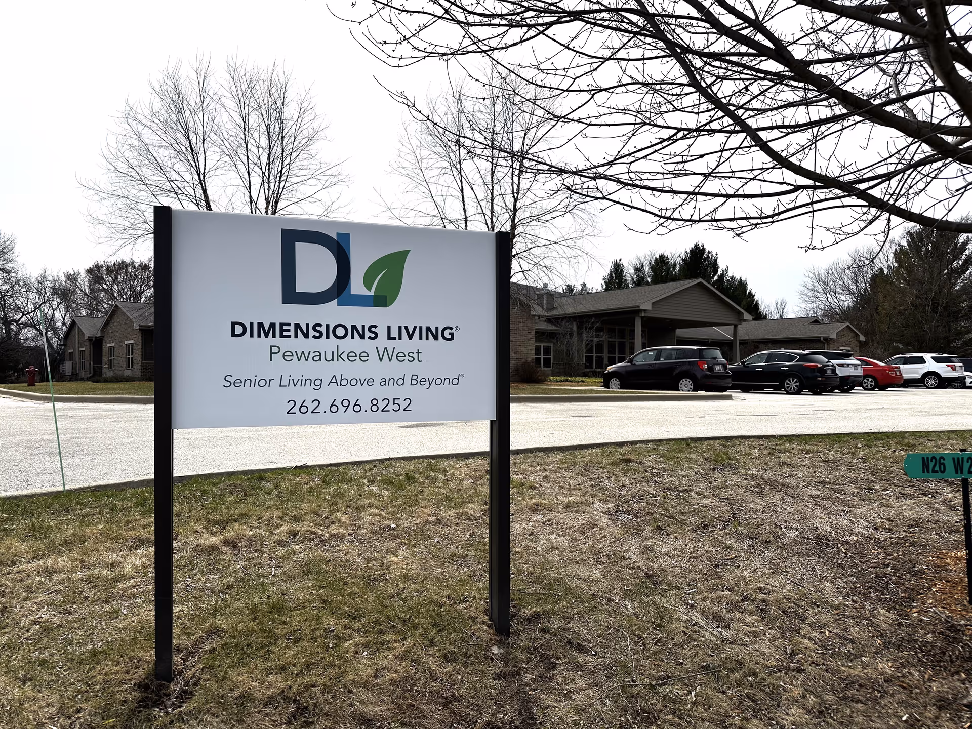 Outdoor view of the Dimensions Living Pewaukee West senior living facility sign with the building and parked cars in the background. The sign displays the facility name, tagline, and phone number.
