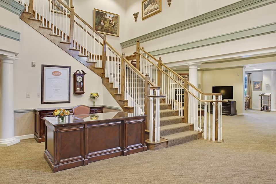 Interior view of a senior living facility lobby area with a wooden reception desk adorned with flower arrangements, a staircase with wooden handrails and white balusters leading to an upper floor, a wall clock, framed artwork, and a TV in the background.