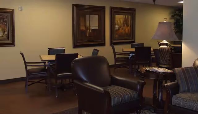 Interior view of a senior living facility common area with dark leather armchairs, a side table with a lamp, several wooden tables with chairs, and framed artwork on the walls.