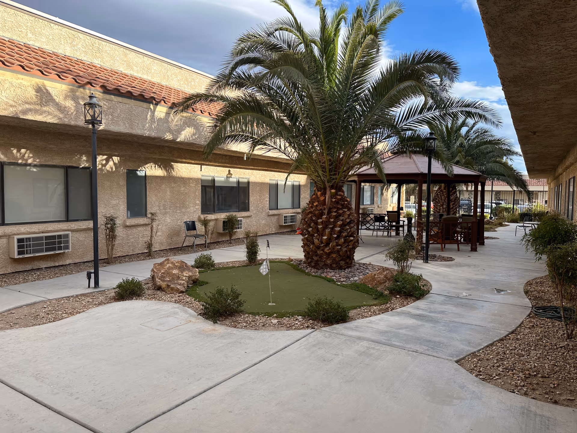 Outdoor courtyard area at Mission Pines Nursing & Rehabilitation Center featuring a putting green with a small flag, palm trees, a gazebo with seating, and a paved walkway surrounded by desert landscaping and building walls with windows and air conditioning units.
