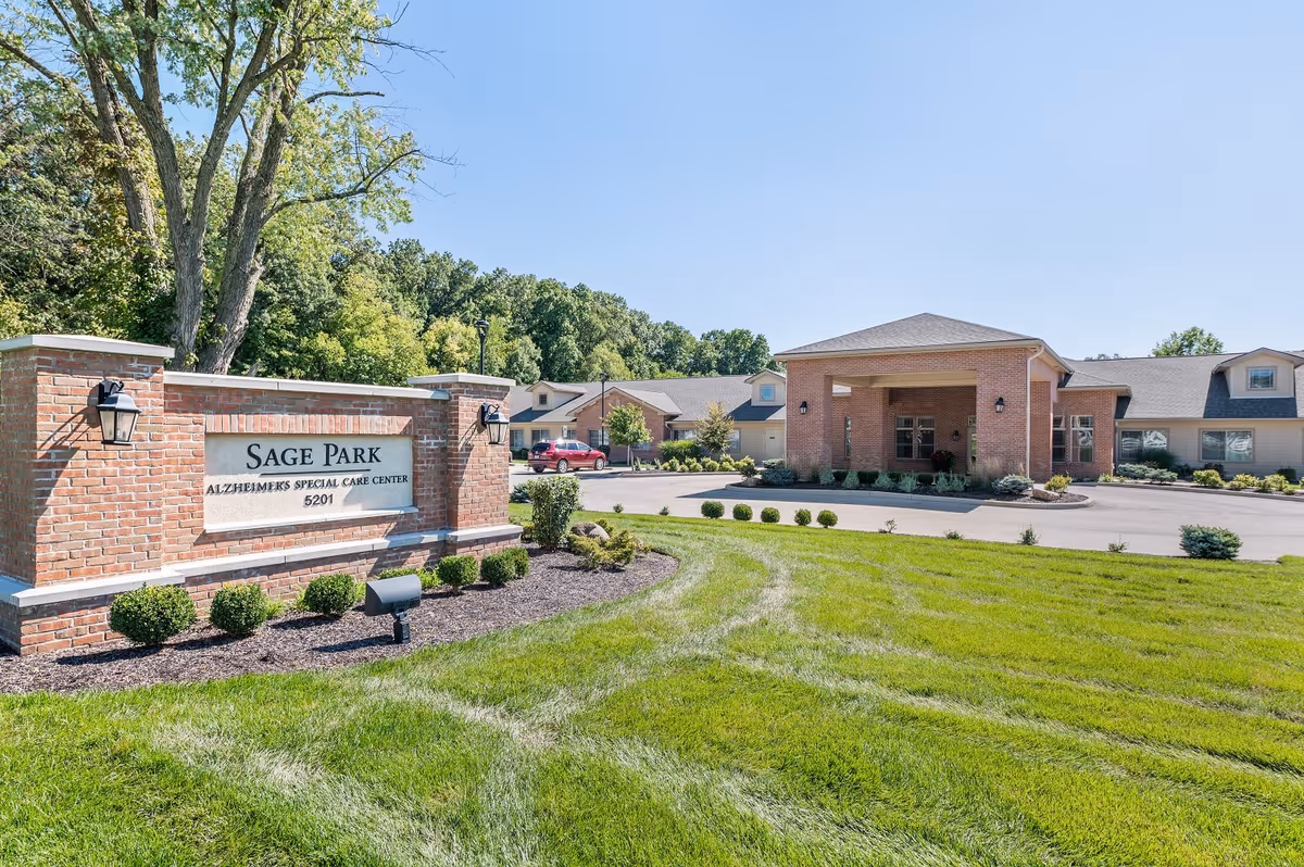 Exterior view of Sage Park Assisted Living & Memory Care facility showing a brick entrance sign with the name and address, a well-maintained lawn, shrubs, trees, and the front entrance of the building under a clear blue sky.