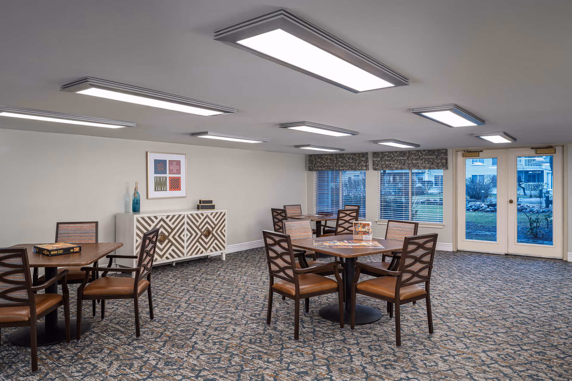A well-lit common area with multiple square tables and chairs arranged on a patterned carpet. There is a decorative cabinet against the wall with a framed artwork and a vase on top. Large windows and glass doors provide a view of an outdoor garden area.