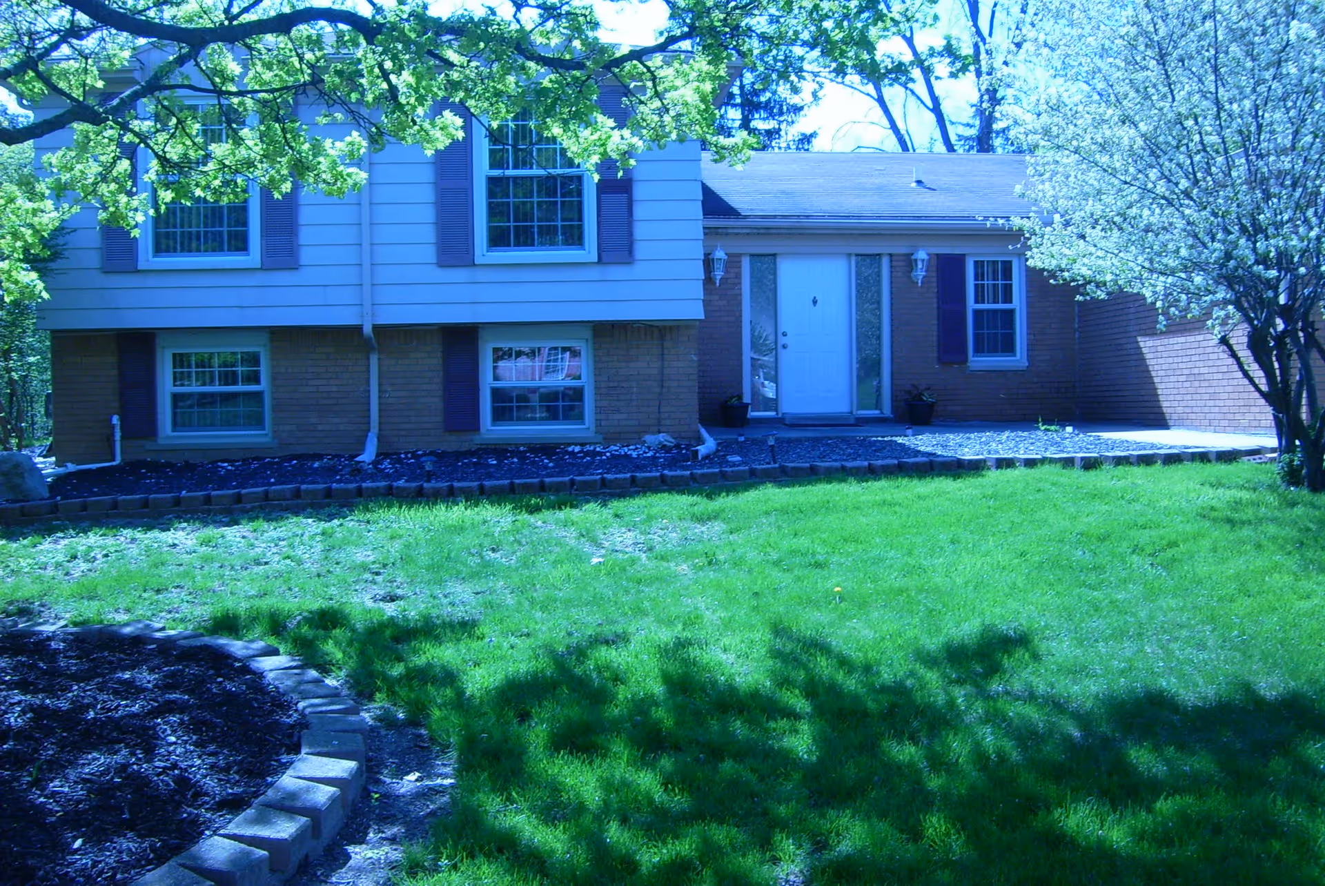 Front exterior view of a two-story house with a white door, multiple windows with shutters, a well-maintained green lawn, and trees with green leaves and white blossoms.