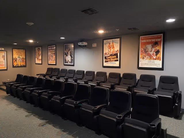 A small movie theater room with two rows of black reclining seats arranged in front of a gray wall. The wall is decorated with framed vintage movie posters. The room has dim lighting and a projector mounted on the ceiling.