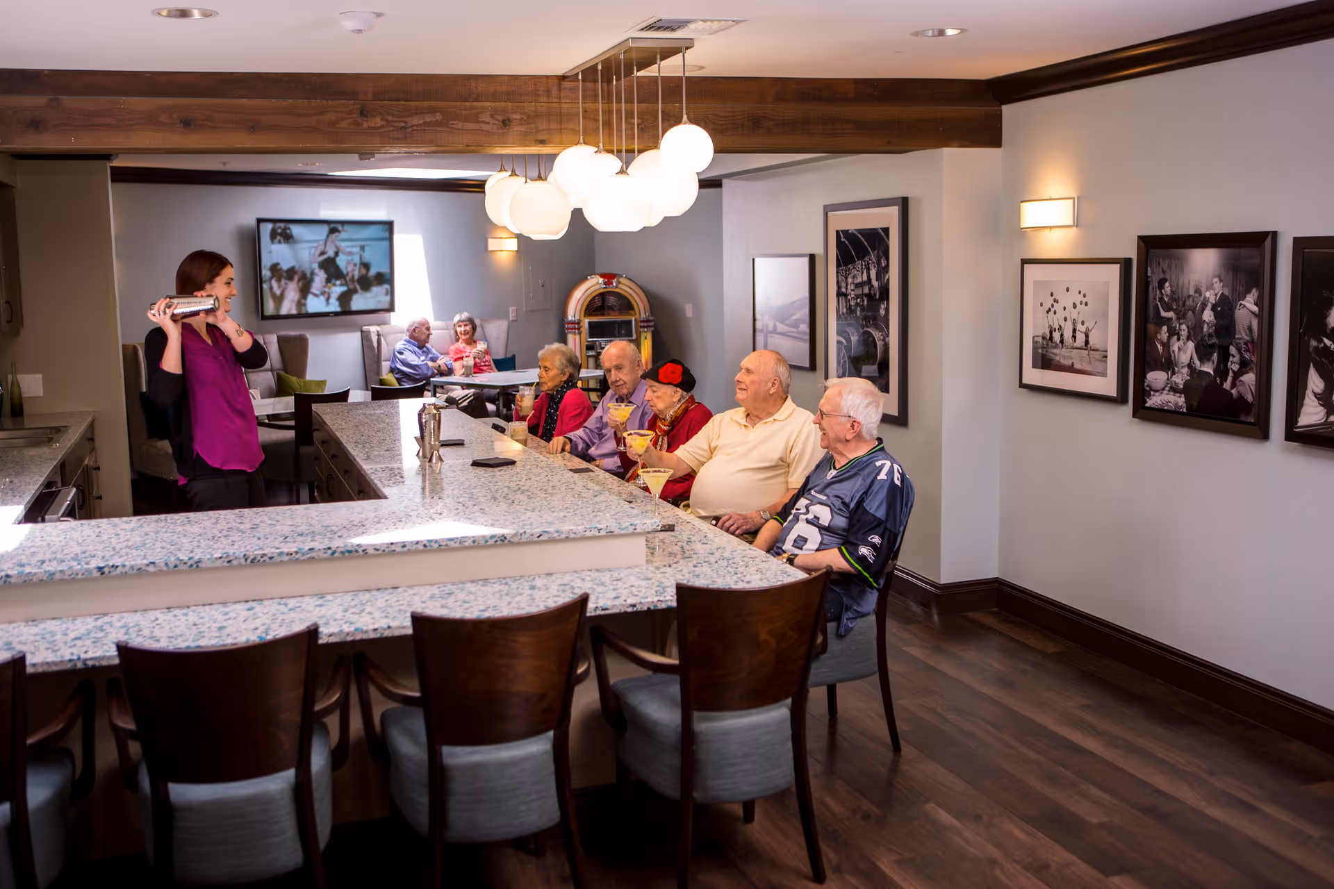 A group of elderly people sitting at a bar counter in a senior living facility, enjoying drinks while a woman behind the counter shakes a cocktail shaker. The room has modern decor with framed black and white photos on the wall, a jukebox in the background, and a television mounted on the far wall.