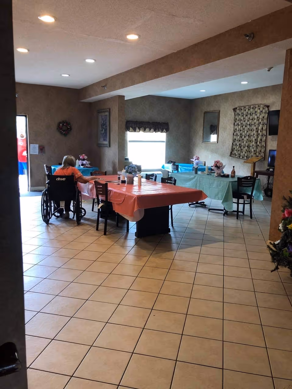 A dining area in a senior living facility with several tables covered in colorful tablecloths. A person in a wheelchair is seated at one of the tables. The room has tiled floors, beige walls, framed artwork, and a window with a valance. There are flowers and condiments on the tables.