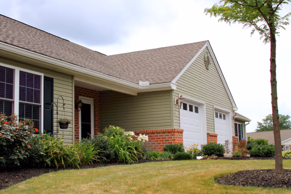 Exterior view of a single-story residential building with light green siding and brick accents. The building features two white garage doors, a small covered porch with a door, windows with dark shutters, and a well-maintained garden with various plants and flowers in front. A tree is visible on the right side of the image.
