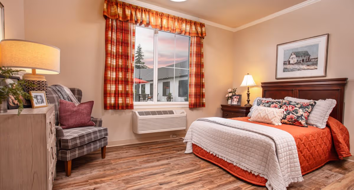 A cozy bedroom with a wooden bed frame, an orange bedspread, and multiple decorative pillows. There is a wooden nightstand with a lamp and framed photos beside the bed. A plaid armchair with a red pillow sits next to a wooden dresser with a table lamp and framed photos. The window has red and orange plaid curtains, and outside the window, part of a building and a tree are visible. The room has wood flooring and beige walls.