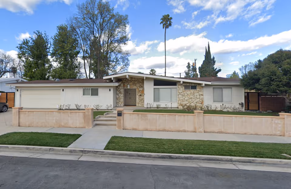 Front exterior view of a single-story residential building with a stone and white panel facade, a two-car garage on the left, a small front yard with trimmed bushes, a low beige wall along the sidewalk, and a clear blue sky with some clouds and tall trees in the background.