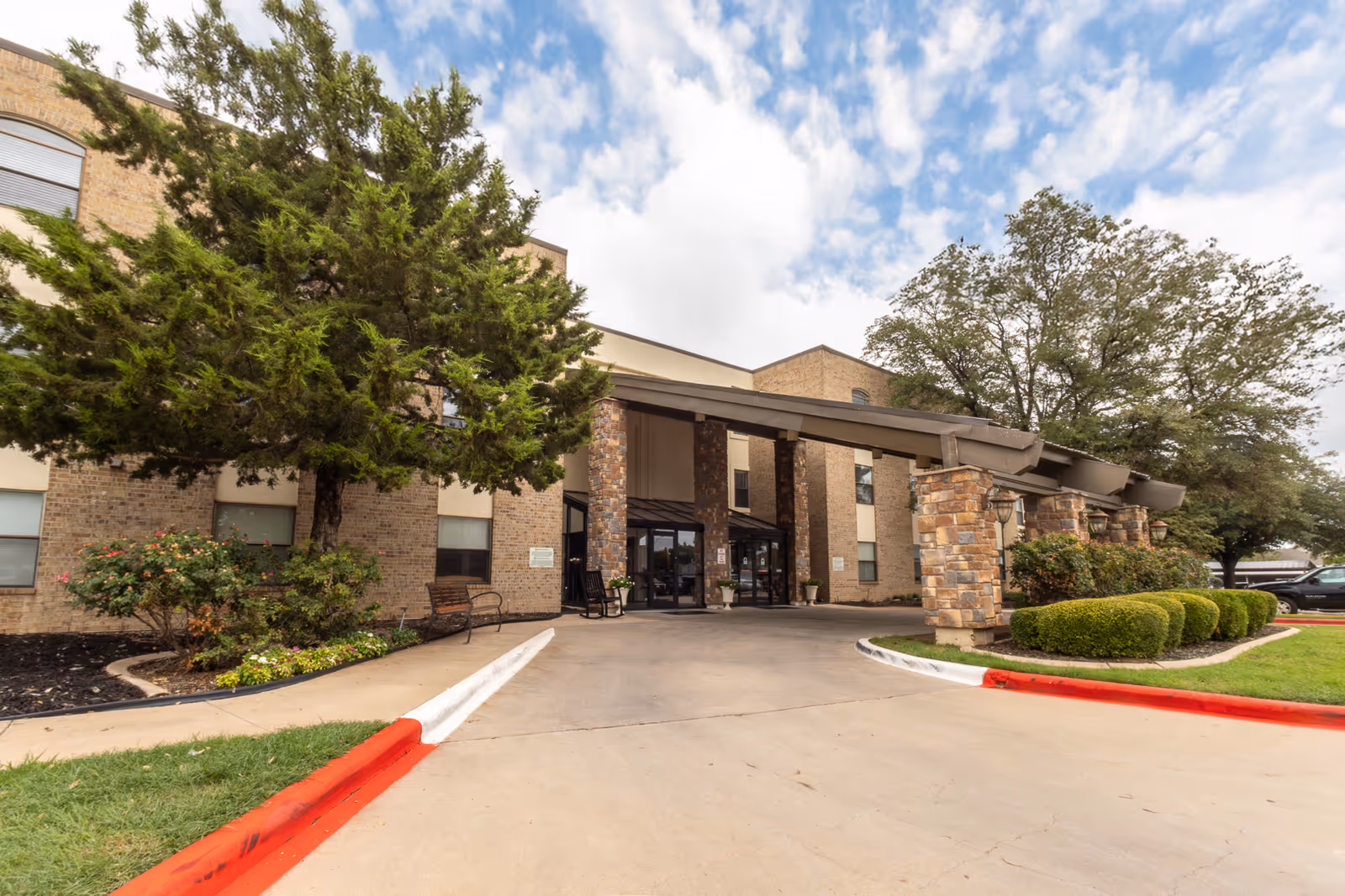 Entrance of a senior living facility named University Place Independent Living, featuring a covered driveway supported by stone pillars, surrounded by landscaped bushes, trees, and a partly cloudy sky.