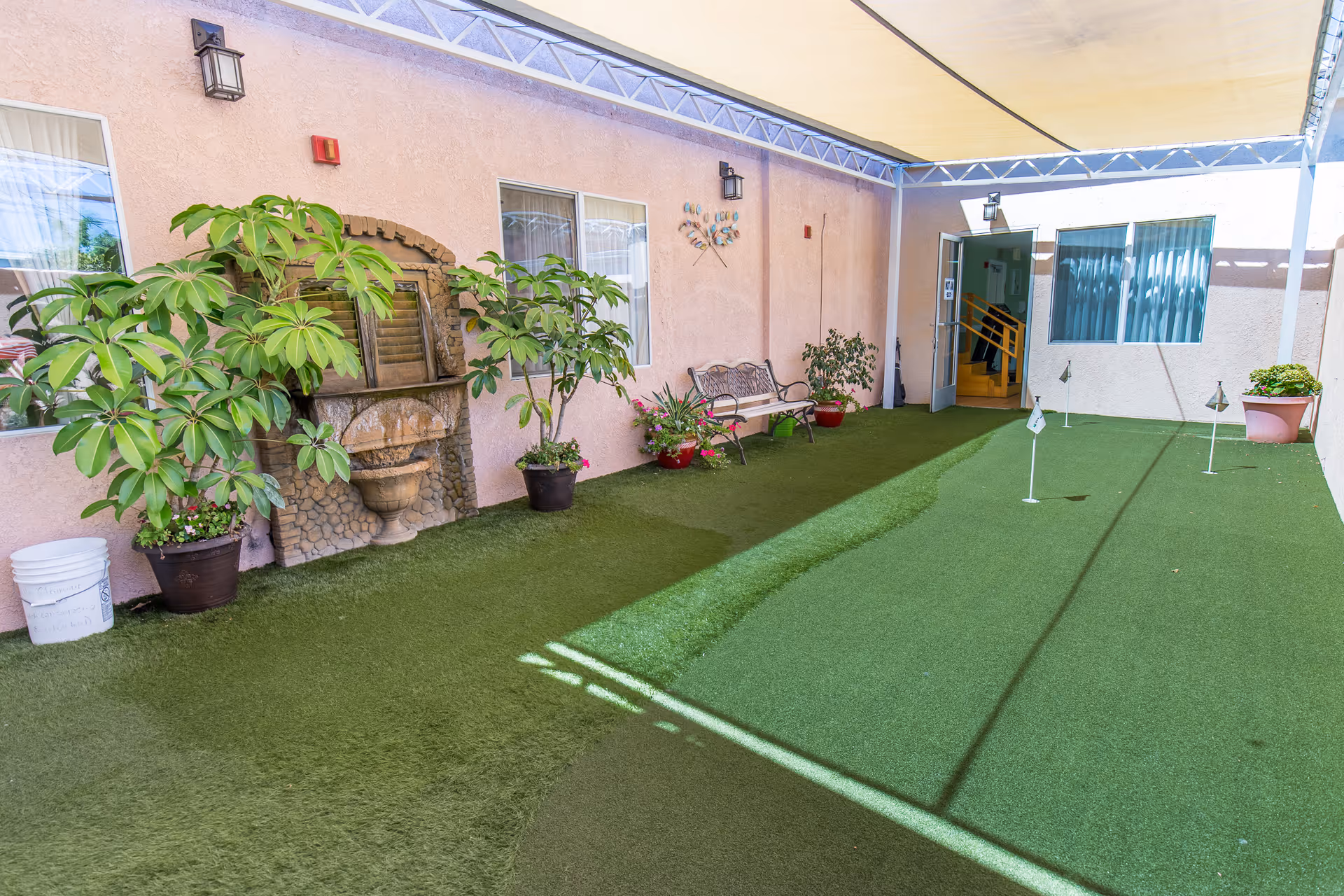 Covered outdoor putting green area with artificial turf, potted plants, a decorative wall fountain, a bench, and an open door leading inside a building.
