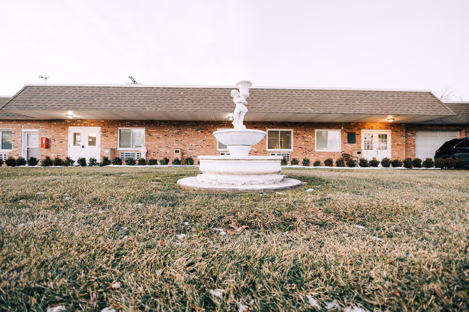 A low-angle view of a white decorative fountain with a cherub statue in the center of a grassy lawn in front of a single-story brick building with multiple windows and doors under a shingled roof.