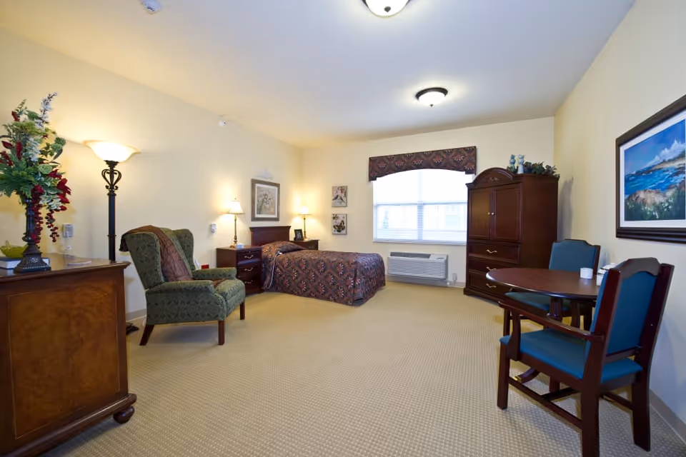 A well-lit bedroom in Avalon Springs Health Campus featuring a single bed with patterned bedding, a green upholstered armchair, a wooden dresser with a floral arrangement, a round wooden table with two blue cushioned chairs, a large wooden armoire, and framed artwork on the walls. The room has beige carpet and cream-colored walls with two ceiling lights and a window with blinds and a valance.