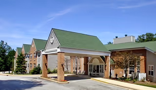Exterior view of Brookdale Wickliffe senior living facility showing a building with a green roof, brick and beige siding, a covered entrance, and a driveway under a clear blue sky.