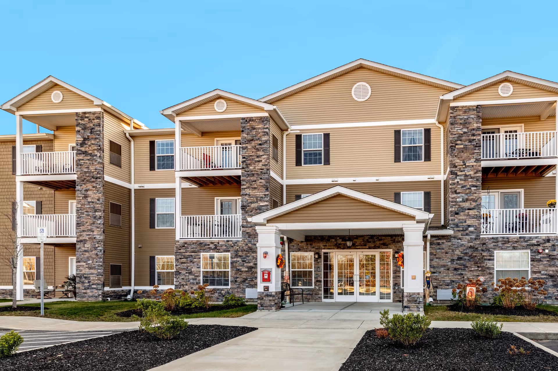 Front exterior view of a three-story residential building with beige siding and stone accents. The building features multiple balconies with white railings and a covered entrance with double glass doors. There are landscaped areas with small bushes and a paved walkway leading to the entrance under a clear blue sky.