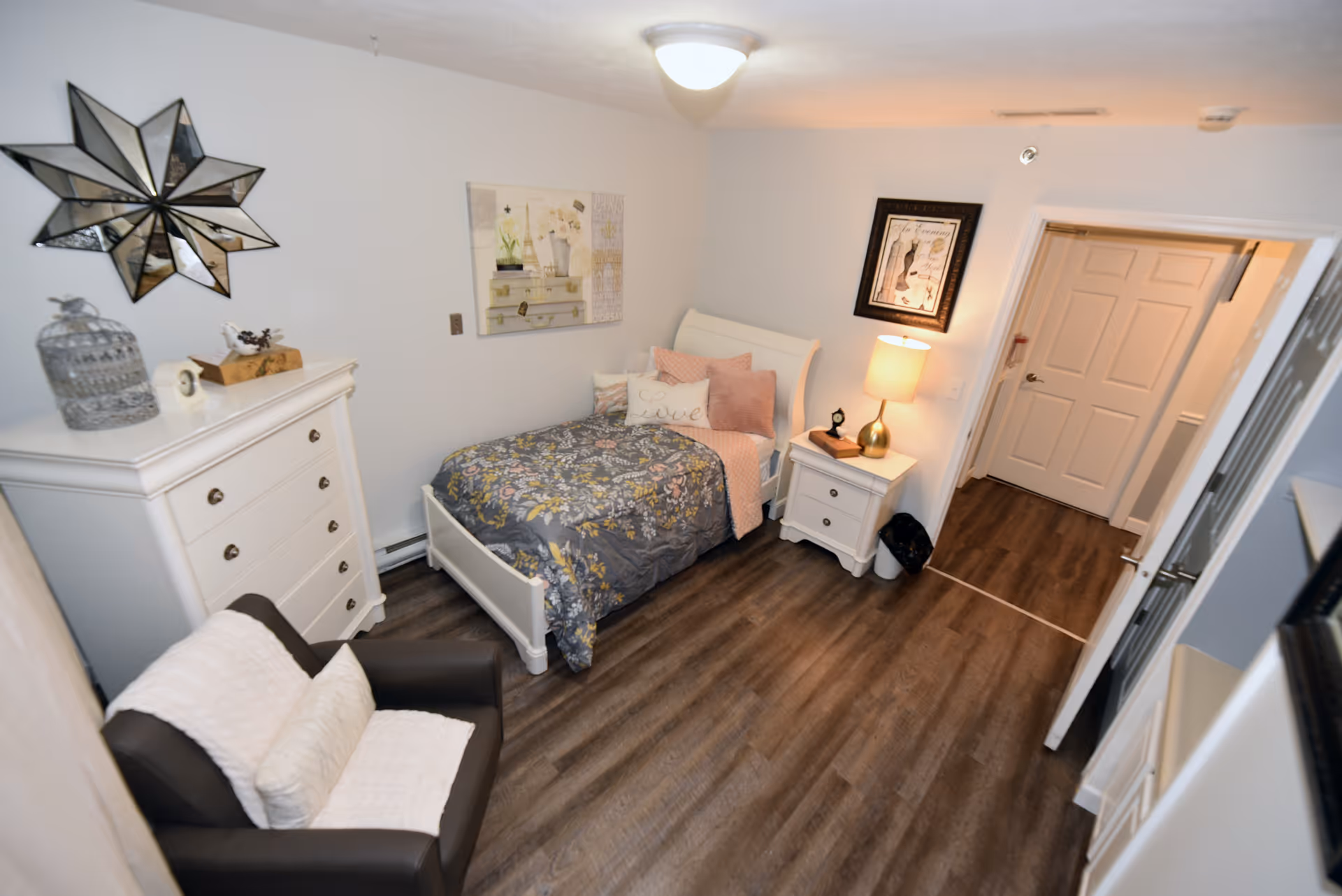A cozy bedroom with a single bed covered in a floral comforter and several pillows, including one with the word 'Love'. Next to the bed is a white nightstand with a lamp and framed artwork above it. Across from the bed is a white dresser with decorative items on top. A comfortable armchair with cushions is positioned near the dresser. The room has wood flooring and white walls with additional wall art and a ceiling light fixture.