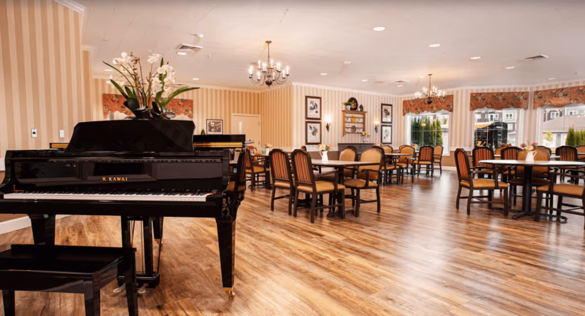 A spacious dining room with wooden floors, multiple tables and chairs arranged neatly, a black grand piano with a floral arrangement on top, striped wallpaper, framed artwork on the walls, and large windows with floral valances letting in natural light.