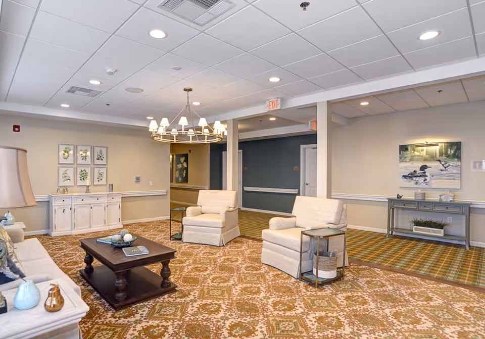 A cozy living room area in Ashton Commons featuring two beige armchairs, a wooden coffee table with decorative items, a white sideboard with framed botanical prints above it, a console table with a painting of ducks on the wall, and a patterned carpet. The ceiling has recessed lighting and a chandelier.