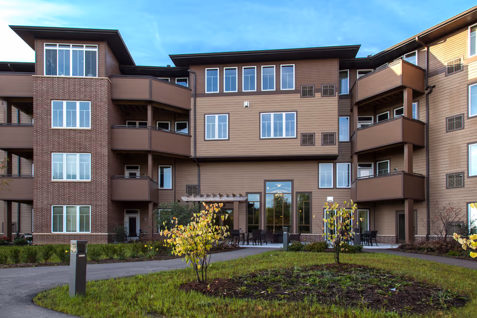 Exterior view of a multi-story senior living facility building with brown siding and brick accents. The building features balconies, large windows, and a small landscaped area with young trees and grass in the foreground under a clear blue sky.