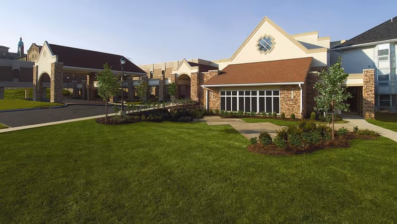 Exterior view of Marymount Place Assisted Living facility featuring a well-maintained lawn, landscaped garden beds, and a building with stone and beige siding, large windows, and a covered entrance.