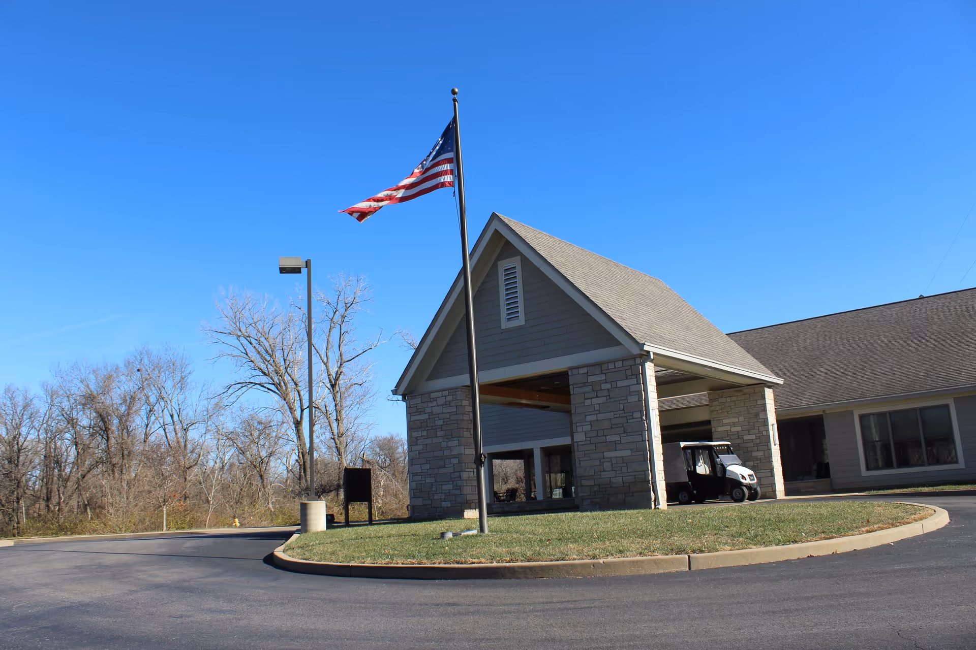 Exterior view of Sherbrooke Village Living Center showing the entrance with a covered driveway, an American flag on a flagpole, a golf cart parked under the entrance canopy, and leafless trees in the background under a clear blue sky.