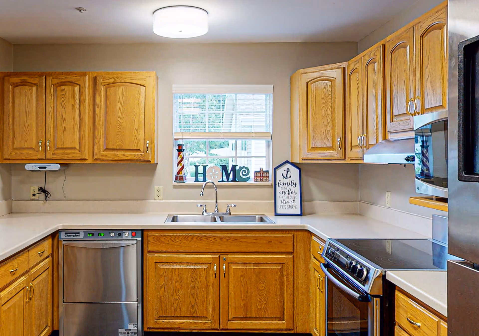 A kitchen with wooden cabinets, a stainless steel dishwasher, a double sink under a window with decorative items spelling HOME, a stove with a glass cooktop, a microwave, and a refrigerator. The walls are beige and there is a round ceiling light fixture.