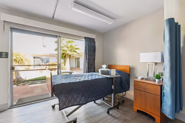 A single bed with a dark blue quilt in a healthcare facility room. The bed has an overbed table with a basket and some items on it. Next to the bed is a wooden nightstand with a lamp, a small plant, and a phone. The room has a large sliding glass door that opens to an outdoor courtyard with palm trees and other plants. There is a blue privacy curtain partially visible on the right side.