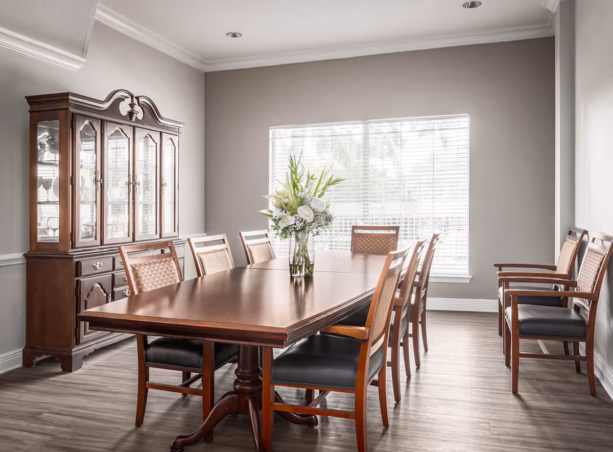 A bright dining room with a large wooden dining table surrounded by eight wooden chairs with cushioned seats. A vase with white and green flowers is placed in the center of the table. Against the left wall, there is a wooden china cabinet with glass doors displaying glassware. The room has light gray walls, a large window with white blinds, and wood flooring.
