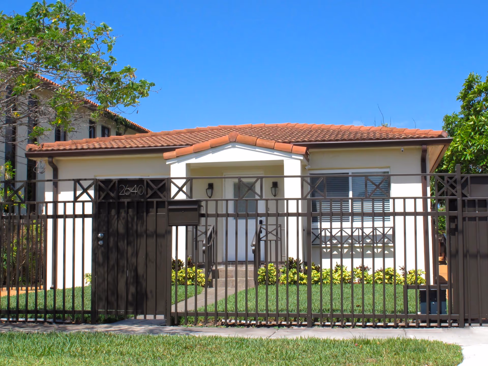 Front exterior view of a single-story house with a tiled roof, white walls, and a black metal fence with a gate. The house has a small porch with steps leading to the front door, flanked by two wall-mounted lights. There is a green lawn and some shrubs in front of the house under a clear blue sky.