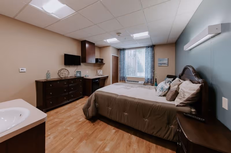 A cozy bedroom in a senior living facility with a large bed covered in beige and brown bedding, a wooden headboard, and multiple pillows. The room features a wooden dresser with decorative items, a small wall-mounted TV, a kitchenette area with a microwave, and a window with sheer and patterned curtains allowing natural light to enter. The floor is wooden, and the walls are painted in beige and blue tones.