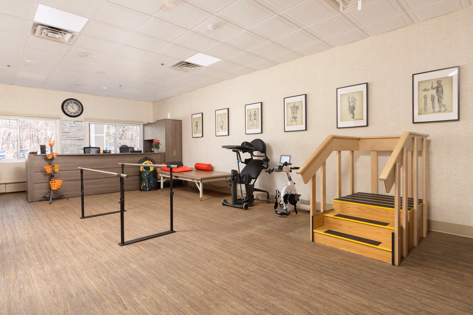 A physical therapy room with wooden flooring and beige walls. The room contains parallel bars for walking exercises, a stationary exercise bike, a padded therapy table with red bolsters, and a small wooden staircase with handrails. There is a reception desk with office chairs and a clock on the wall above it. Several framed anatomical drawings are hung on the wall.