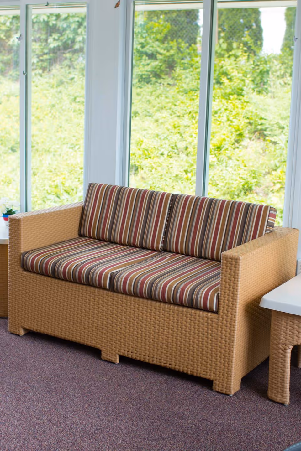 A wicker loveseat with striped cushions in shades of brown, red, and beige, placed indoors near large windows showing green foliage outside.