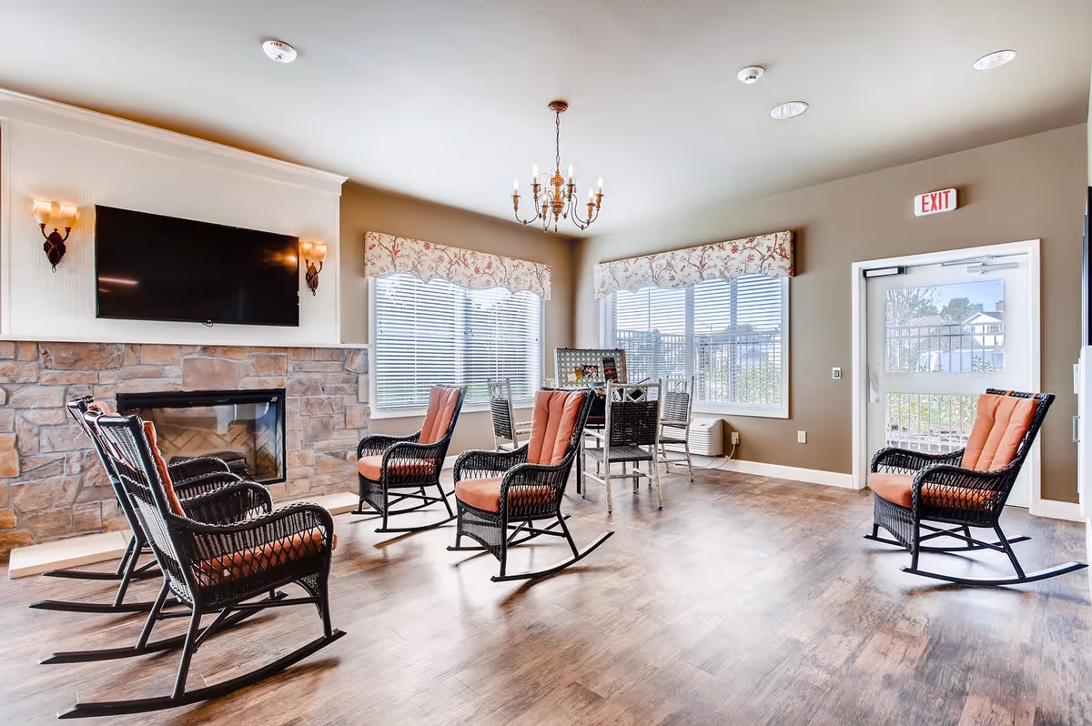 A bright and spacious common area with six black wicker rocking chairs with orange cushions arranged around a stone fireplace with a mounted flat-screen TV above it. The room has large windows with floral valances, a chandelier hanging from the ceiling, and a small table with chairs near the windows. There is a door with an exit sign above it leading outside.