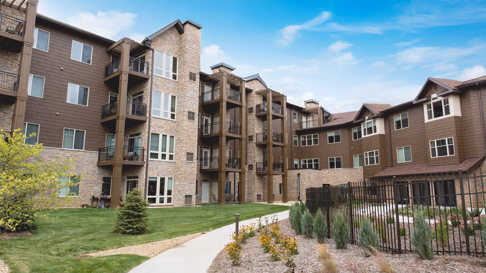 Exterior view of a multi-story senior living facility building with brown siding and stone accents. The building features balconies and numerous windows. In the foreground, there is a well-maintained lawn, a curved concrete walkway, and landscaped areas with small trees, shrubs, and flowers under a partly cloudy sky.