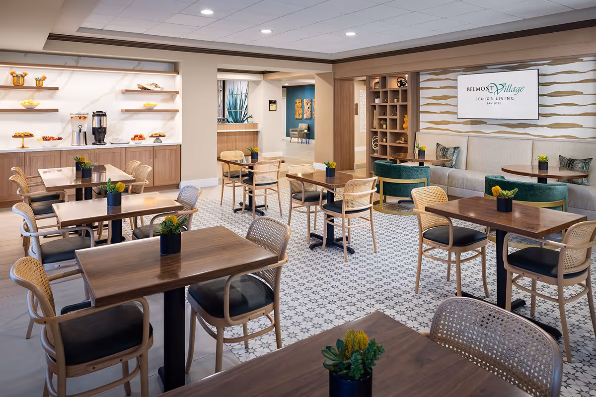 A bright and modern dining area in Belmont Village Senior Living San Jose featuring multiple wooden tables with black cushioned chairs. The floor has a patterned tile design, and there are small potted plants on each table. The back wall has shelves with decorative items and a beverage station with coffee and snacks. A large sign on the wall displays the Belmont Village Senior Living San Jose logo. The space is well-lit with recessed ceiling lights and has a cozy seating area with green velvet chairs and a beige cushioned bench.